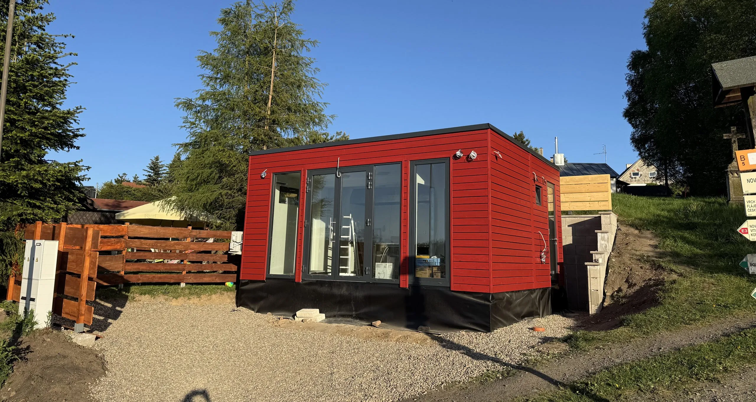 Rozestavěné bistro — kompaktní objekt s červeným dřevěným obkladem a prosklenou stěnou, uvnitř stojí štafle, u paty stavby je vidět hydroizolace a čerstvý štěrk.